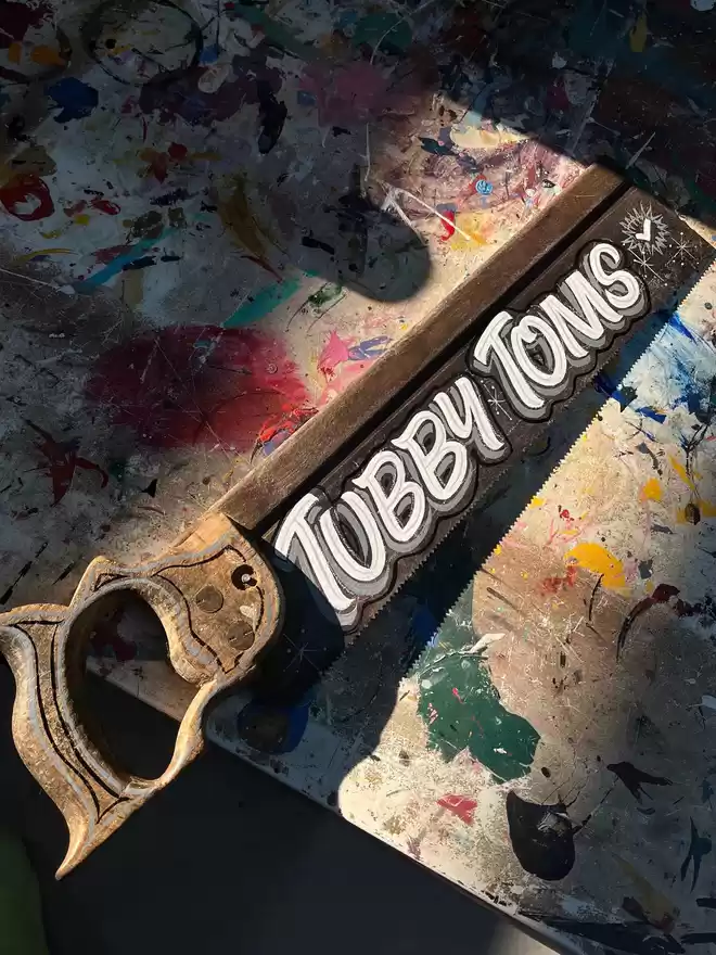 Hand-painted vintage saw spelling 'Tubby Toms' with white, grey and black lettering in a casual style