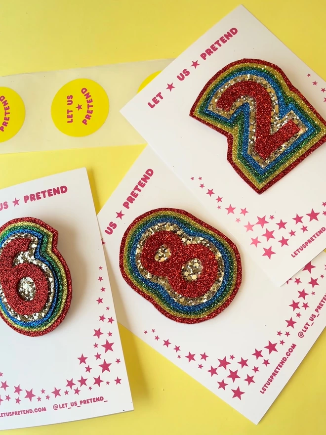 Three rainbow glitter badges arranged against a yellow backdrop. They are in the ages 'six', 'eight' and 'two'. The centre number is red with a border of five layers of colour. The colours are chunky gold glitter, blue, green, gold, and red.