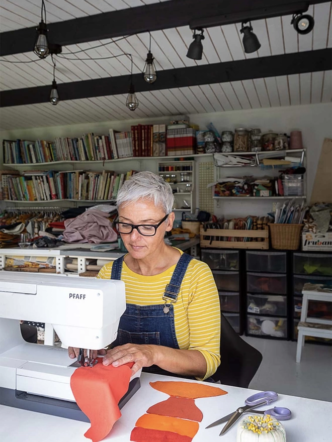 Manda McGrory wearing elbow top and blue dungarees with glasses sewing an orange toy lobster on a sewing machine in her studio
