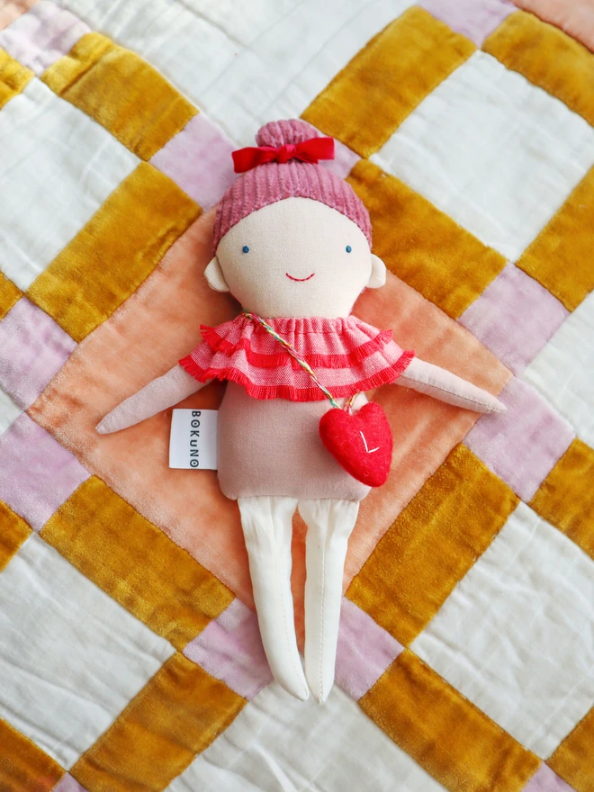 Handmade cloth doll with a pink corduroy hair with a red velvet bow and a layered red ruffle collar and pink outfit, wearing a small red felt heart bag with the letter “L”