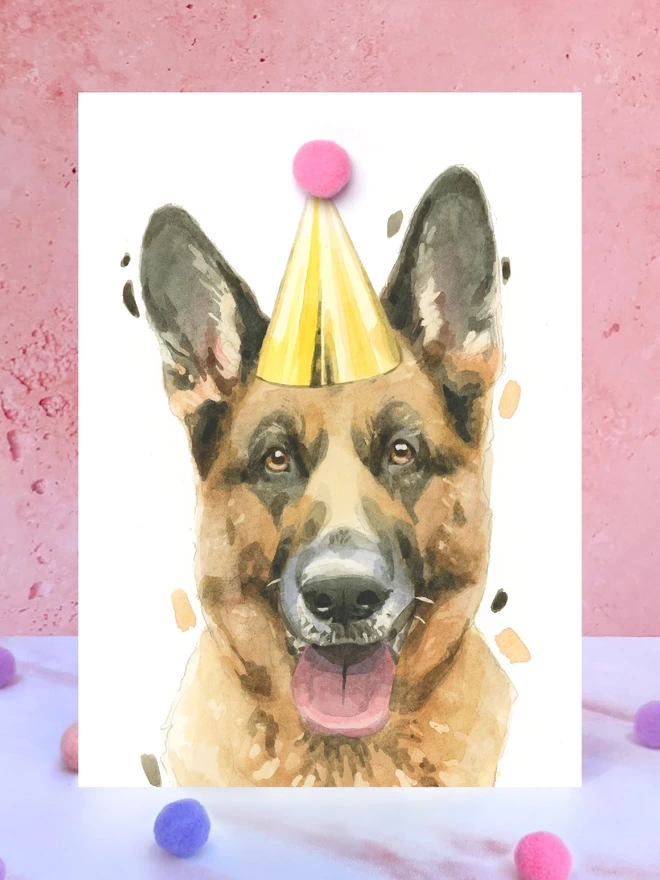 A greeting card featuring a hand painted design of a german shepherd, stood upright on a marble surface surrounded by pompoms. 