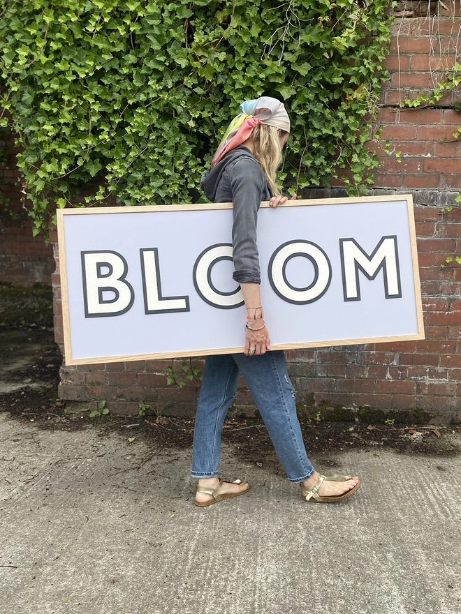A person standing in front of greenery holding a painted wooden sign which reads BLOOM on a lilac painted back board 