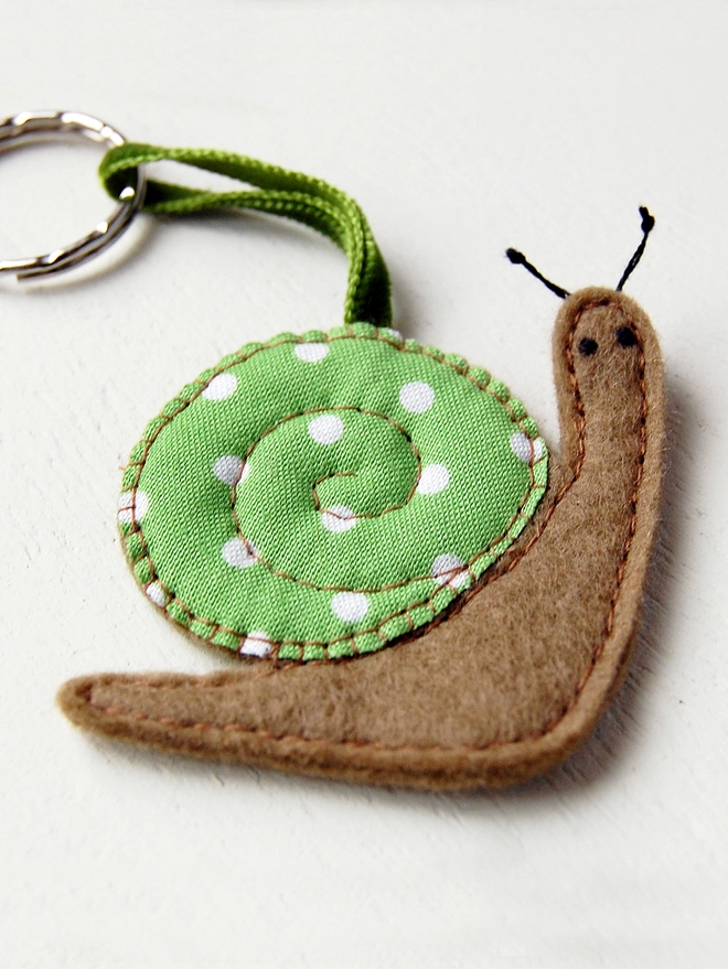 A homemade felt snail keyring with a brown body and a green spotty shell lays on a white desk.