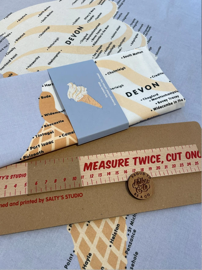 Measure Twice Cut Once wooden ruler, in a cardboard presentation pack, lies on a teatowel with an icecream image, next to another folded up Ice cream design teatowel. A showcase of Saltys Studio Products