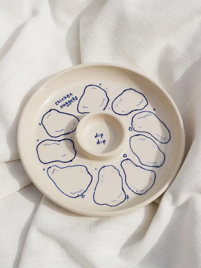 Snack Chicken Nuggets & Dip Plate, a white snack dish sitting on a white bed cloth. It is covered in blue outlines for chicken nuggets. 
