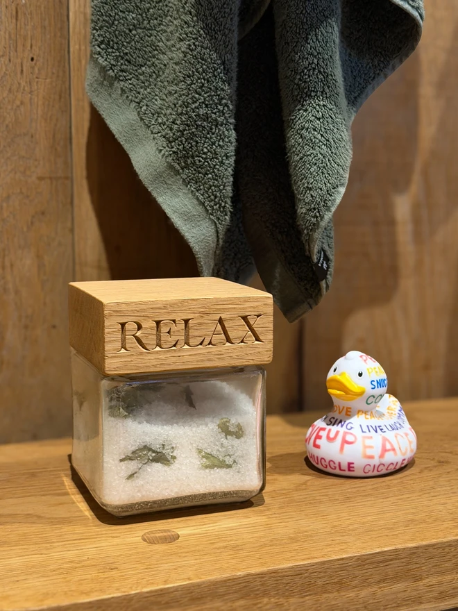 Oak 'relax' jar. A glass jar with an engraved oak lid on a wooden bathroom shelf, with a green towel and a rubber duck.