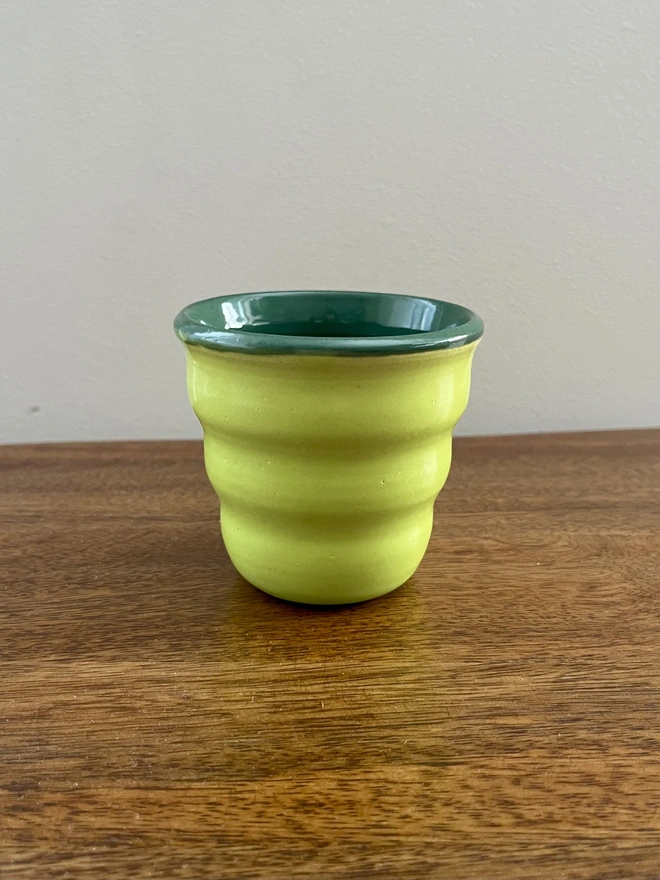 mini ceramic tumbler with wavy sides on a wooden table. the tumbler has a lime green glaze on the outside and forest green on the inside and rim. 