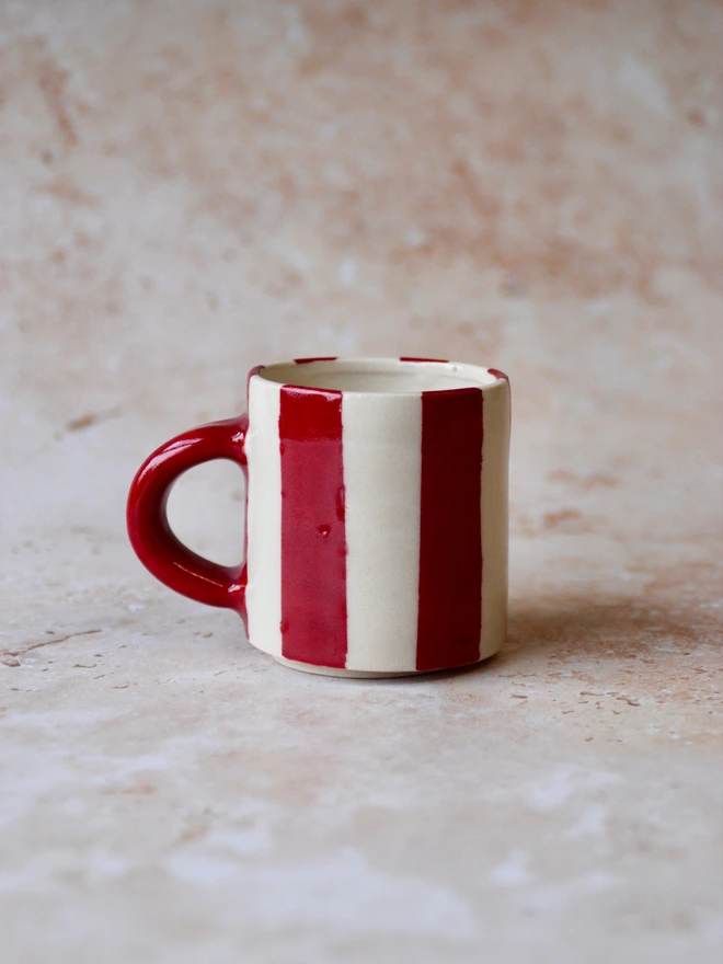 Handmade ceramic mug with bold red and cream vertical stripes and a glossy red handle, photographed on a neutral stone background.