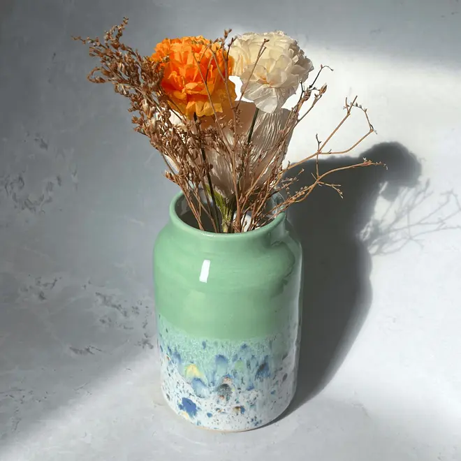 Shoreline Large Vase, a colourful vase placed on a stone surface in front of a plain backdrop. It is accompanied by flowers. 