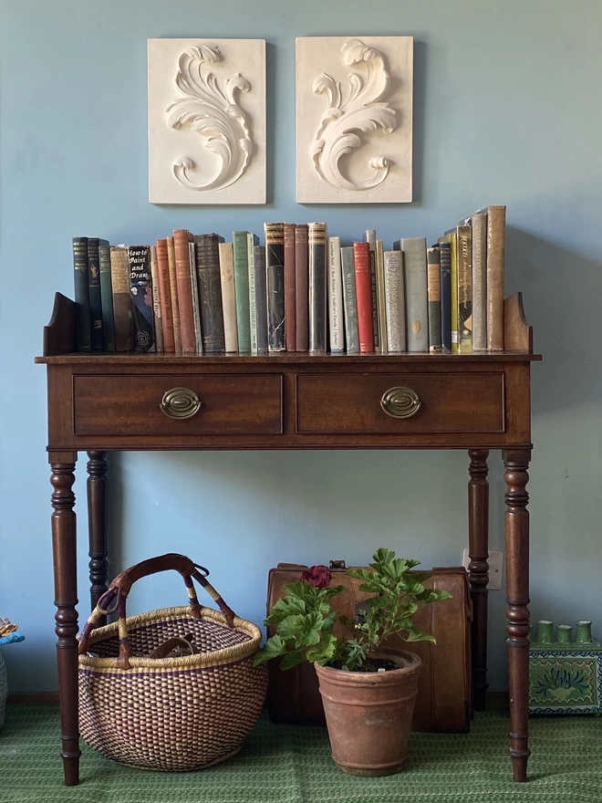 Pair of plaster bas-relief wall plaques with acanthus leaf design and table and bookshelf underneath