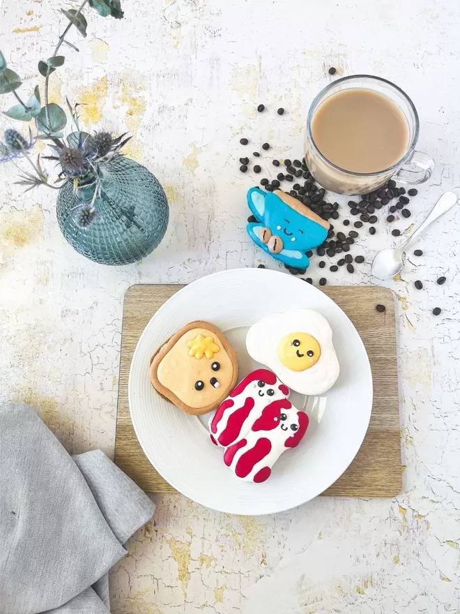 a plate topped with bacon, fried egg & toast shaped macarons and a cup of coffee with a vase of flowers against a cracked yellow table