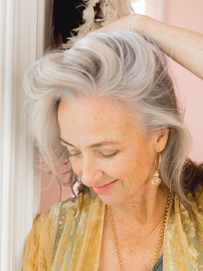 Grey haired woman in yellow top holding her hair back to reveal a large gold hoop earring with a Frida Kahlo mexican heart charm