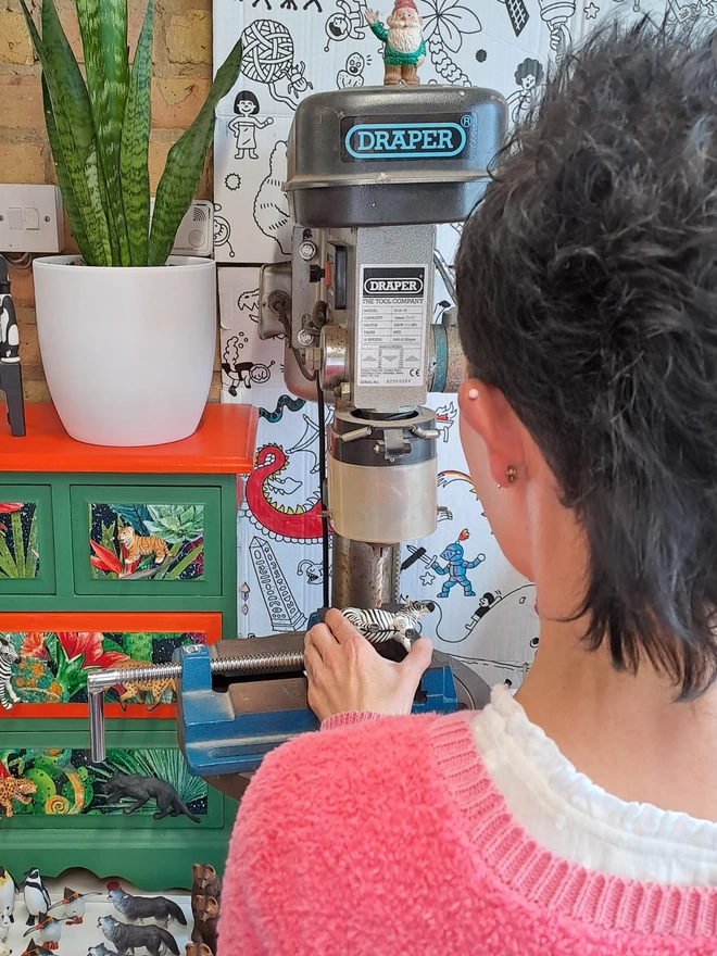 A hand holds a plastic zebra on a clamp on the table of a pillar drill. A white tray of plastic animals sits below the pillar drill to the left. There is a small chest of drawers decorated in a brightly coloured jungle theme with animal drawer knobs attached with a snake plant in a white vase sitting on top. The zebra is being drilled and made into an animal drawer knob by Candy Queen Designs.