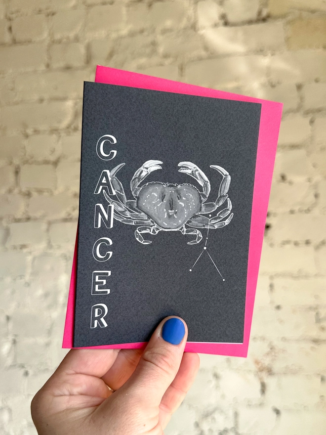 a greetings card featuring a greyscale illustration of a zodiac style crab with the text “CANCER” with a neon pink envelope