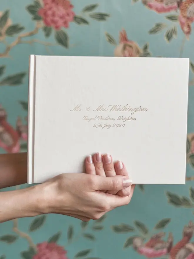Personalised Calligraphy Font Wedding Guest Book, held in front of a floral background. 