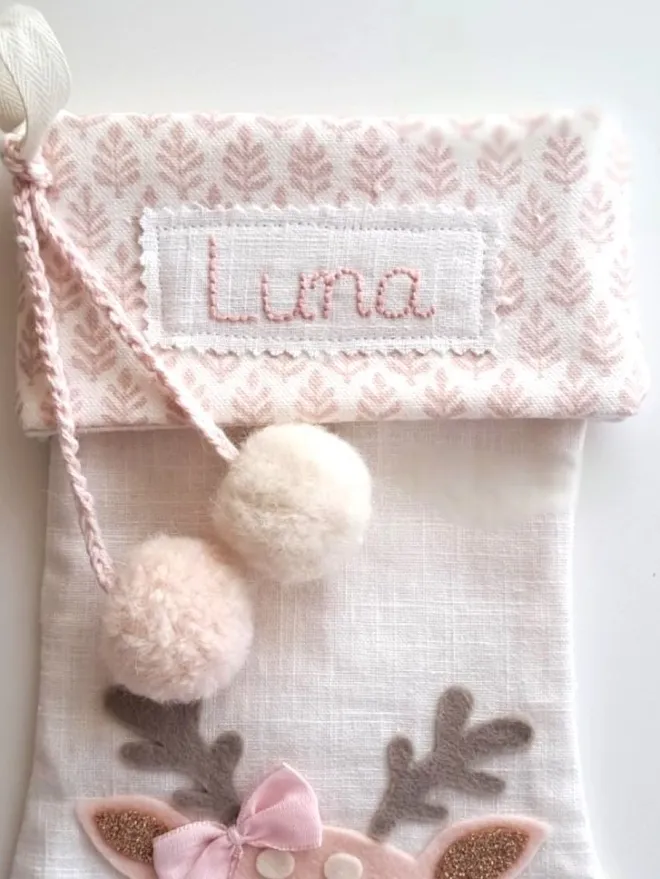 Baby Christmas stocking, close up of pink hand embroidered name for personalisation option, there are two fluffy pompoms at the side