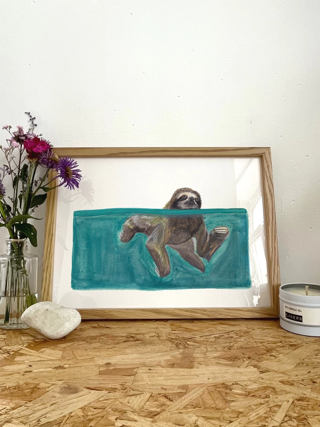 A print of an illustration of a sloth swimming in blue water, in a frame next to some flowers, a rock and a candle