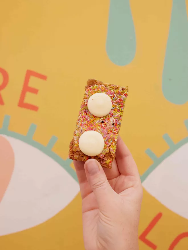 Single slice of Funfetti blondie with sprinkles and white chocolate buttons held in one hand against a colourful background