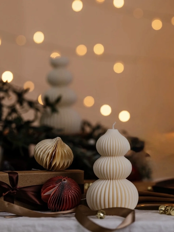 Signature Christmas Tree Sculpture Candle. A cream ribbed Christmas tree candle beside paper honeycomb baubles and a wrapped gift with satin ribbon, set against warm fairy-light bokeh. The minimalist festive candle makes a thoughtful Christmas gift or stocking filler.