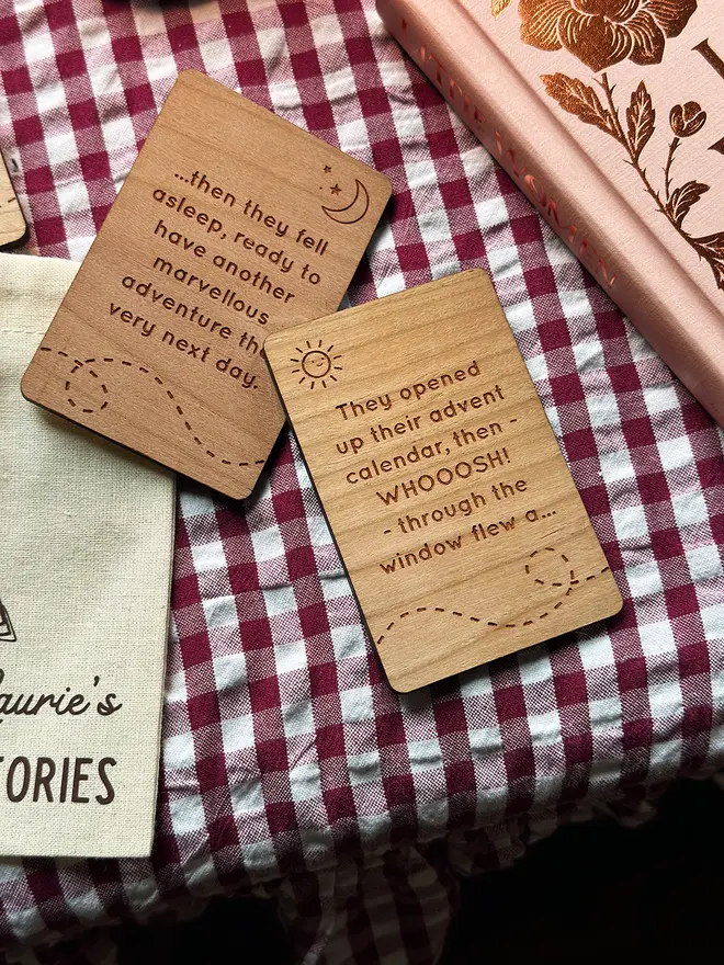 Close-up of engraved wooden Christmas story prompt cards on a red gingham cloth beside a pink book, showing a mix of story beginnings and endings for children’s festive storytelling.