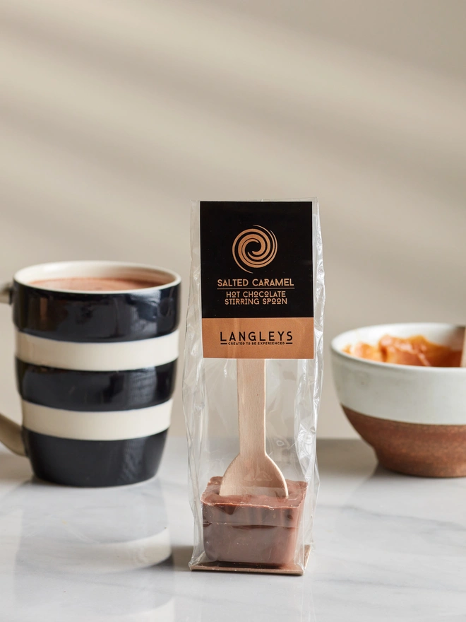 Milk Hot Chocolate Salted Caramel Stirring Spoon, a chocolate serving spoon pictured on a marble table top next to a hot chocolate and a bowl of food. 