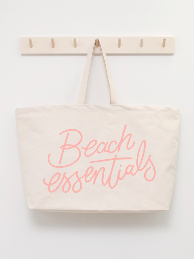 oversized natural canvas tote bag with beach essentials slogan hanging on a wooden hook