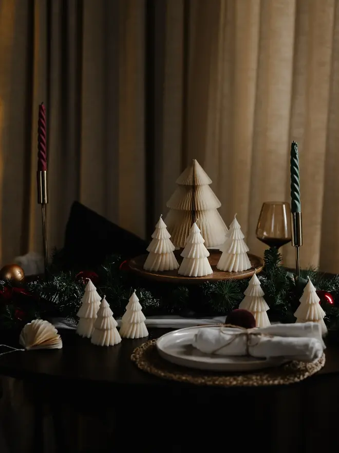 White Christmas Tree Warm Candles (Set Of 2). Cream Christmas tree candles with pleated, layered tiers grouped on a wooden board around a paper honeycomb tree, with pine garland and a twisted green taper candle. The minimalist sculptural candles make an elegant, understated festive centrepiece.