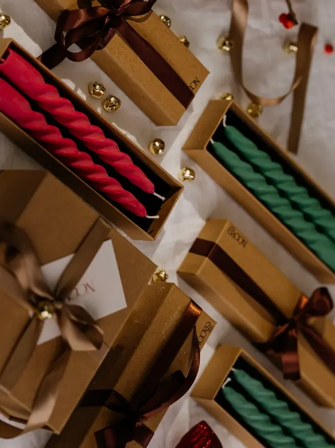 Christmas Spiral Candles Gift Box (Set Of 2). Pairs of red and green twisted taper candles in open kraft Noord gift boxes, surrounded by boxes tied with brown satin ribbon, gold jingle bells and a linen cloth. The gift-ready spiral candles make a festive present or a colourful finishing touch for Christmas tables.