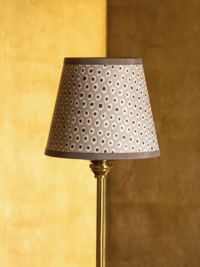 Candle Clip Shades. A small candle lampshade in a soft lilac with cream and plum spots and a matching trim, fitted to a slim brass lamp. The delicate block-printed shade suits a bedside table, hallway or dressing table.