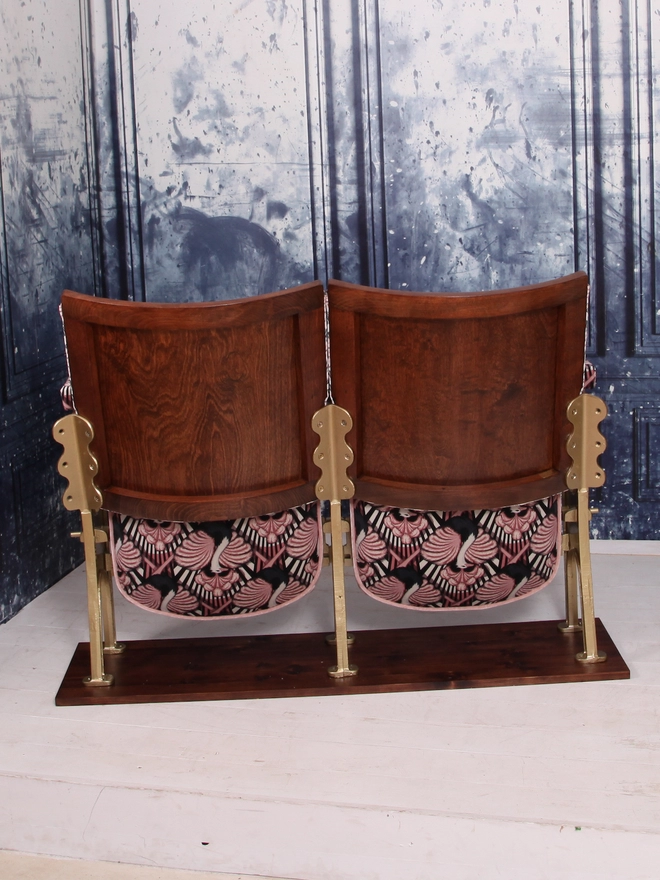 Rear view of a set of two vintage cinema seats upholstered in a pink ostrich Art Deco design velvet with pink panel and piping against a blue marbled wall.  Seats are down