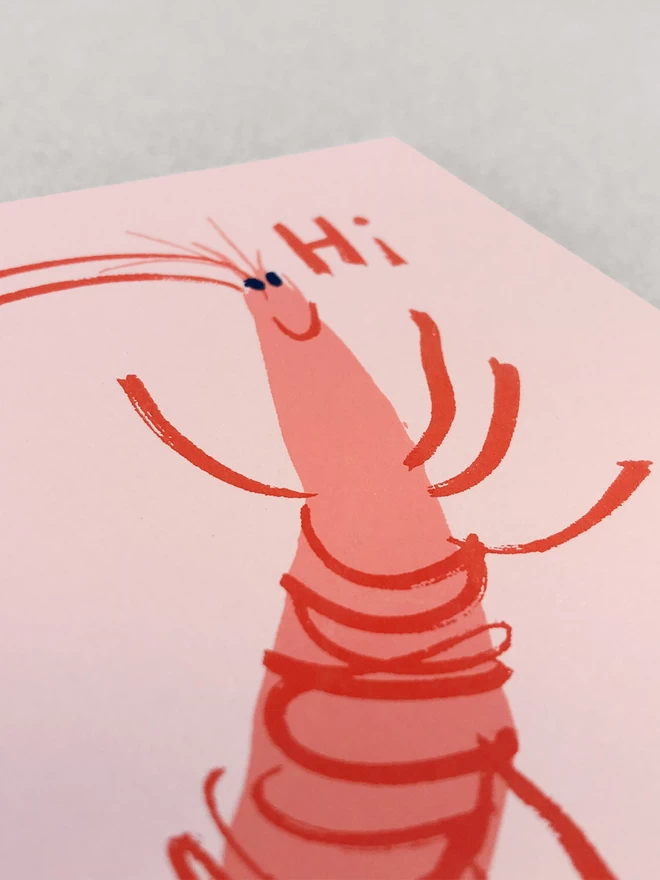 Close up detail of happy printed prawn with Hi written next to his smiling face. Showing a close up of the texture of the pink card, laid on a grey background.