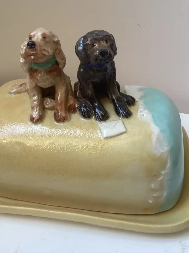 One brown and one black dog sitting on a hand-painted butter dish that looks like a beach which a shoreline.
