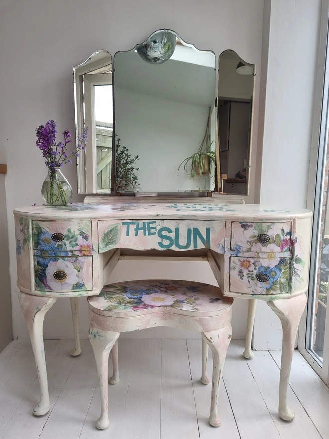 A painted dressing table in white and pale pink with large florals in blues and mauves and text in turquoise that reads 'turn your face to the sun'. The whole piece is distressed in the shabby chic style.