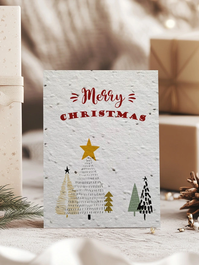 Christmas Card featuring Christmas trees and 'Merry Christmas' across the top. In the background is presents, pine cones and greenery.