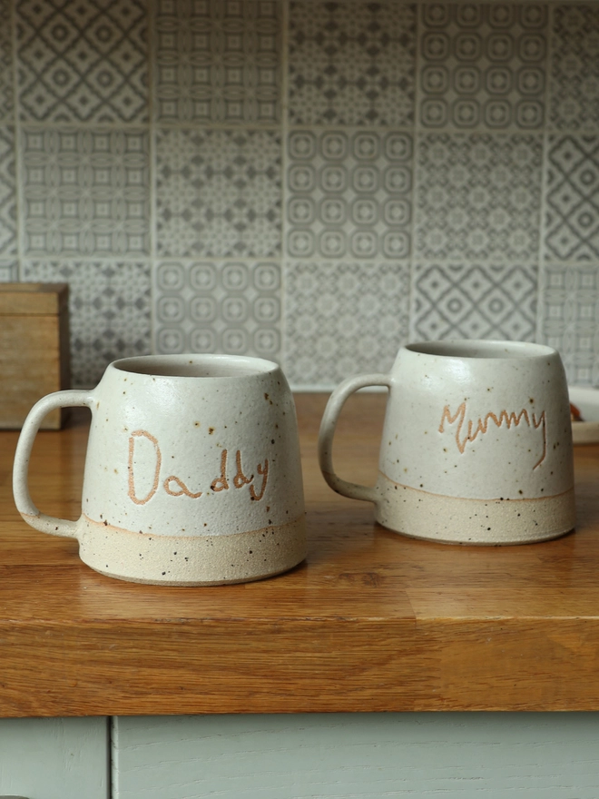 2 Personalised Kids' Handwriting Handmade Stoneware Mugs on a wooden kitchen counter featuring the names Daddy and Mummy 