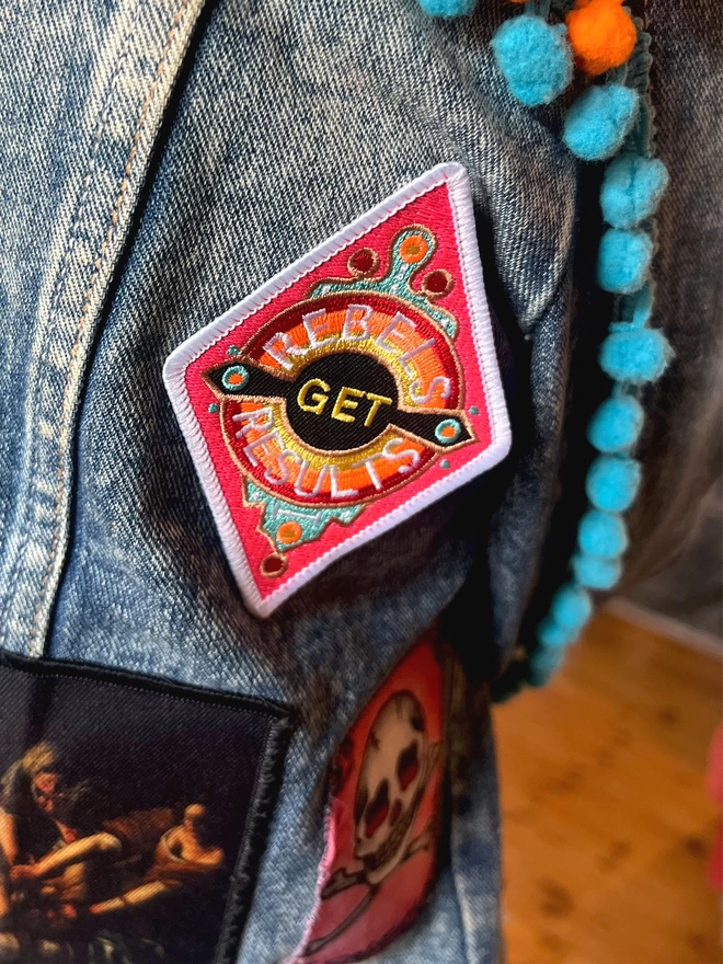 The patch is on a denim jacket with blue and orange trim detail and other patches. The patch is a diamond shape embroidered patch with “Rebels Get Results” at the centre. The design features pink, white, red, yellow and black colours and has a white border.