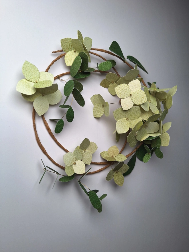 Green hydrangea flower wreath, handmade from paper. There is a cluster of 16 hydrangea blooms on the wreath, which is also adorned with single flowers and two types of dark green leafy stems.