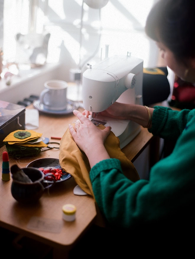 A dark haired white woman sits at a desk using a sewing machine 