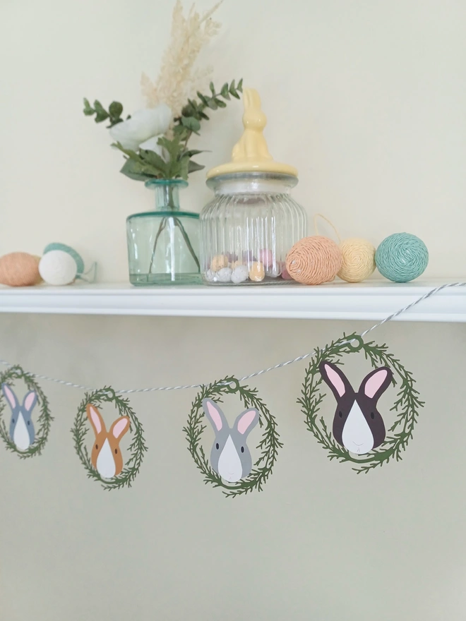 5 Bunny Easter Paper Garland hung from white shelf. Each decoration features a different coloured rabbit face surrounded by an olive green foliage oval card wreath 