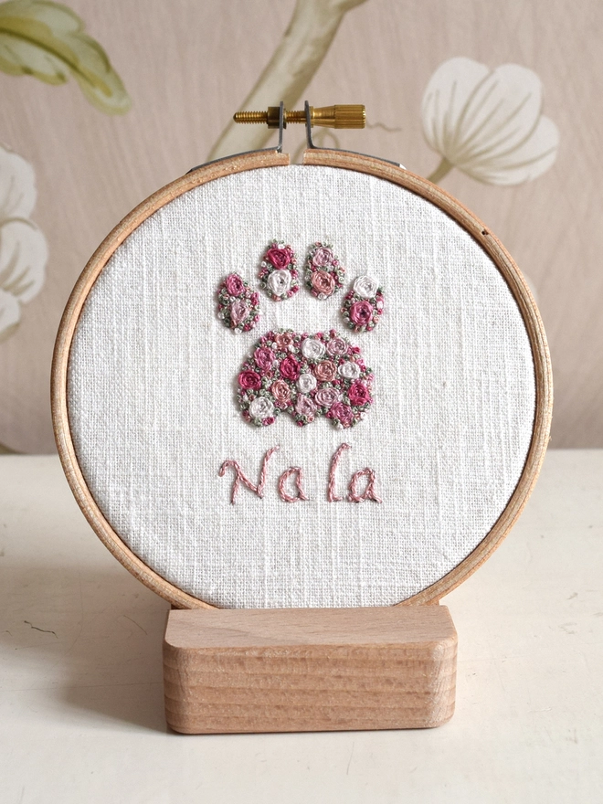 Pink Roses Cat Paw, an embroidery design of Woven Roses and scattered French Knot blossoms in 5 shades of pink with 2 shades of green grass French Knots.  Displayed in an embroidery hoop on a wooden stand.