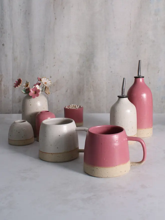 A selection of fuchsia and off-white handmade ceramic pieces including mugs, small and large oil pourers, fuchsia match pot and our vases in mini, small and medium sizes. The medium vase hold an off-white and fuchsia ceramic flower bouquet with gold lustre detailing.