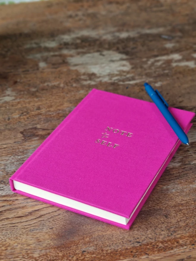 A bright pink notebook printed with the words 'Note to Self' in gold lettering rests on a wooden table top.