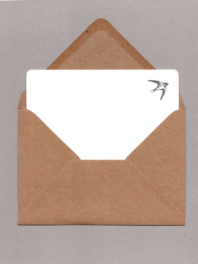 White notecard with a black swallow at the top-right corner inside an open kraft envelope