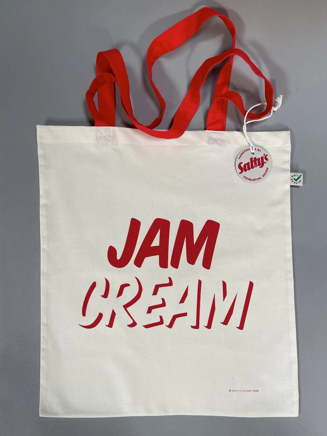  Flat lay of a white, natural coloured tote bag laid down with the word JAM written on top of CREAM, screenprinted in red ink. A Saltys swing tag is tied to one strap. The handles are red.