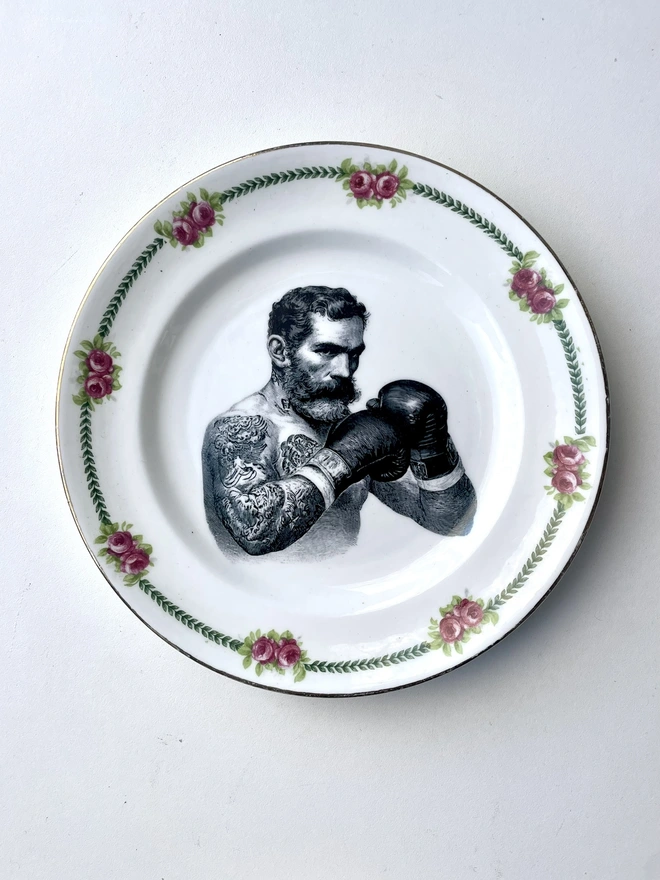 vintage plate hanging on a white wall, it has a decorative border, with a printed vintage illustration of a wild beard east end boxer in the middle 