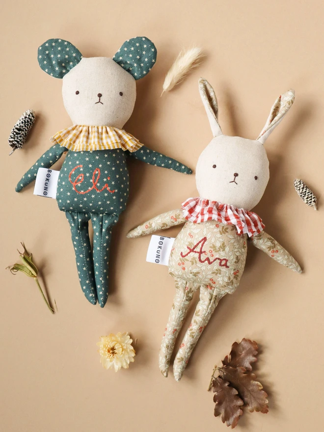 Handmade mouse doll with teal fabric featuring small white stars, a yellow gingham ruffle collar, and the name ‘Eli’ embroidered in red on its body. A white tag with the brand name ‘BOKUNO’ is sewn on the side. Handmade rabbit doll with light floral-patterned fabric, a red-and-white gingham ruffle collar, and the name ‘Ava’ embroidered in red on its body. A white tag with the brand name ‘BOKUNO’ is sewn on the side.