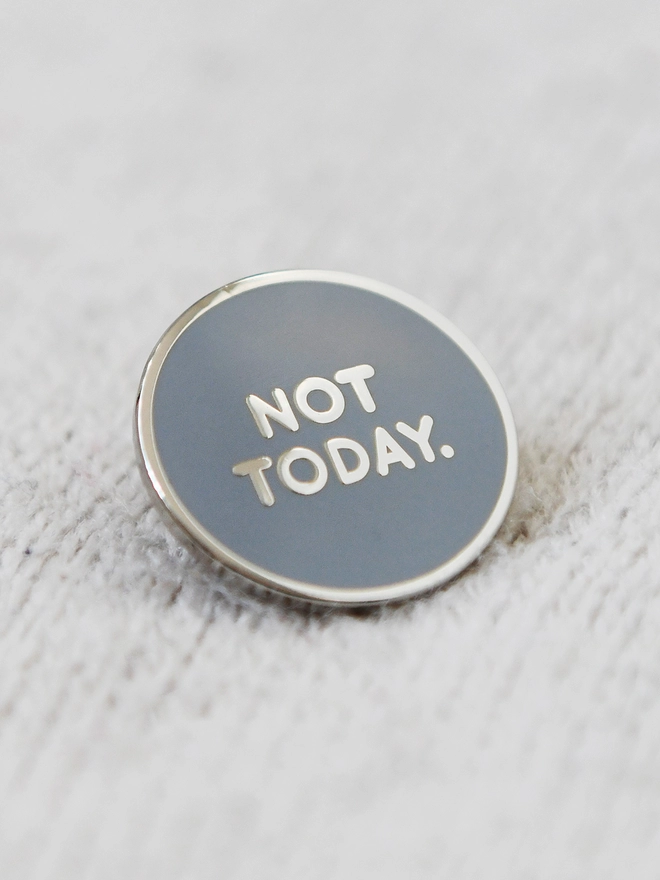 A round grey enamel pin badge with silver words that reads "Not Today" rests on ivory fabric.