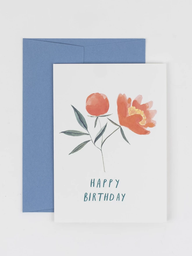 An illustrated greetings card, with corresponding pale blue envelope. The illustration features a peony stem with a closed bud and an open flower with coral petals. There is teal handwritten text underneath that reads "Happy Birthday"