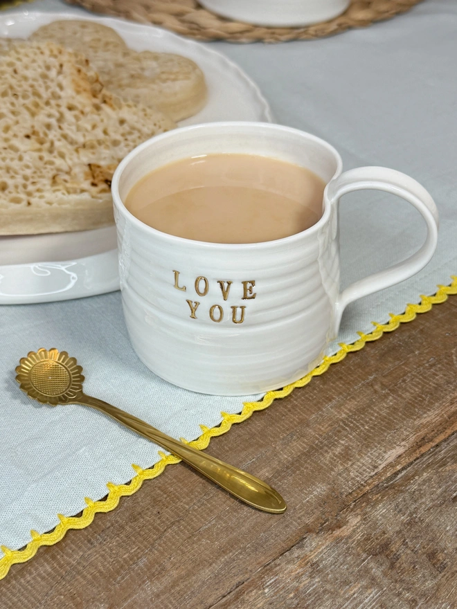 Love You hand thrown porcelain mug with gold lustre text on the side of the mug. The mug sits on a rustic wooden table top with some heart shaped crumpets on a plate and a pretty gold flower shaped tea spoon on a teal table cloth