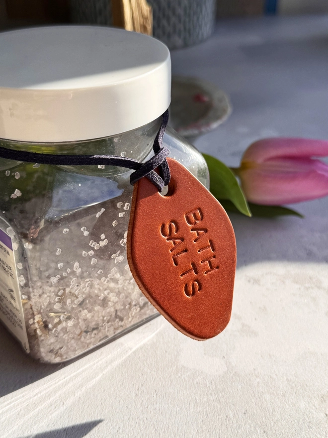 "Three hand-stamped leather tags in brown, cream, and tan, with the words 'RELAX MUMMY,' 'EPSOM SALTS,' and 'BATH SALTS' embossed. Each tag has a cord for tying around jars, displayed on a light, textured surface.